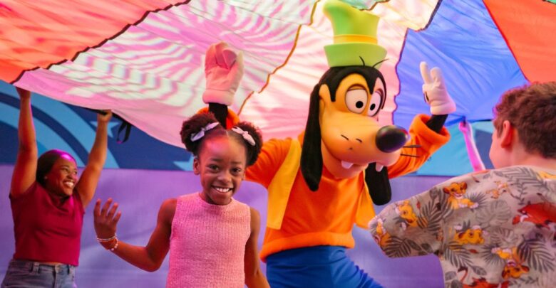 Goofy leads kids and an adult in parachute games at a lively indoor Walt Disney World celebration.