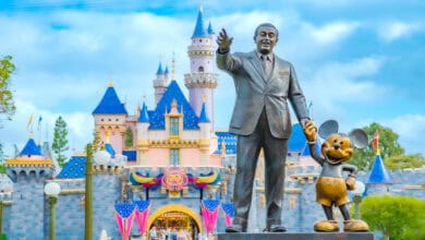 Walt Disney and Mickey Mouse statue welcomes guests in front of the vibrant castle at Disney Resort beneath a bright blue sky.