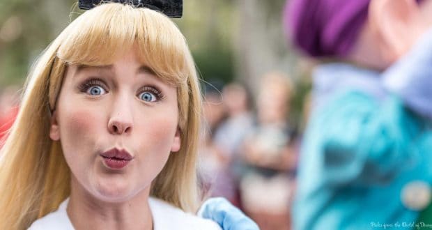 Alice from Alice in Wonderland looks shocked at a Disney park event, reacting to something heard, with guests blurred behind her.