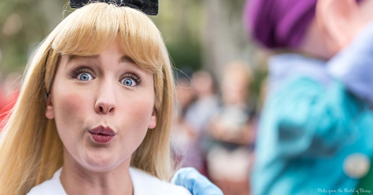 Alice from Alice in Wonderland looks shocked at a Disney park event, reacting to something heard, with guests blurred behind her.