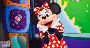 Minnie Mouse beams in her classic red polka-dot dress before vibrant cartoon screens and a globe at Walt Disney World.