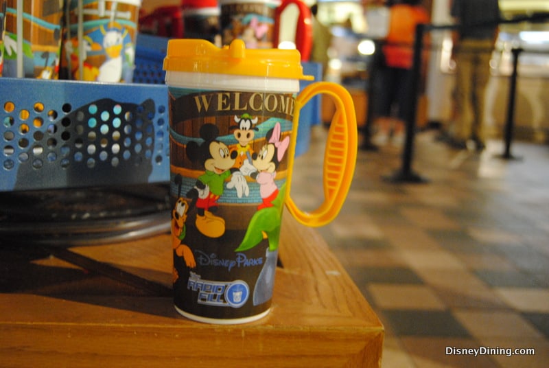 A Disney Parks refillable mug featuring beloved characters and "WELCOME" text rests on a wooden table inside the park.