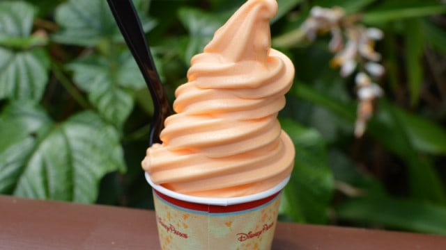 Orange soft-serve in a paper cup with a black spoon, set before lush greenery—a hidden Disney treat as magical as the parks themselves.