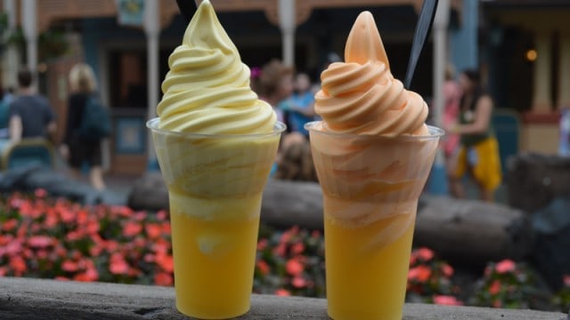 Dole Whip swirled soft serve floats atop pineapple juice at Disney, with blurred park guests and colorful blooms in the backdrop.