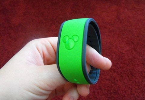 A guest displays a green Disney World FastPass+ wristband featuring the Mickey Mouse logo, with a vivid red backdrop.