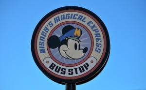 Mickey Mouse in a bus driver hat beams from the "Disney's Magical Express Bus Stop" sign, set against a bright Disney World sky.