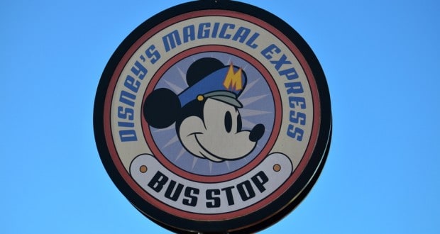 Mickey Mouse in a bus driver hat beams from the "Disney's Magical Express Bus Stop" sign, set against a bright Disney World sky.