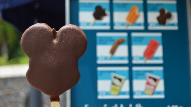 Mickey Mouse ice cream bar, chocolate-dipped, held up in front of a Disney World menu featuring iconic frozen park snacks.