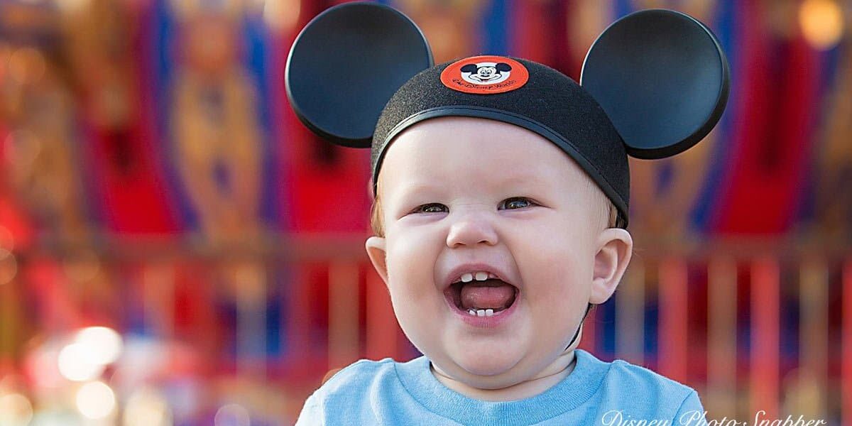 Smiling baby wearing Mickey Mouse ears hat, with an open mouth and visible teeth, in front of a colorful, blurred background.
