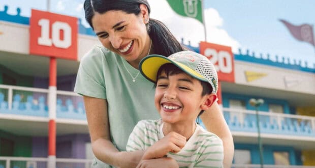 A smiling woman hugs a laughing young boy in a baseball cap outside a colorful building with numbered signs and flags at Hollywood Studios. The clear blue sky adds to their cheerful day—perfect for discovering new Hollywood Studios tips together.