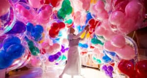 A person in white clothing stands surrounded by large, colorful balloons, creating a vibrant, immersive environment filled with Disney Magic and pink, red, blue, green, and purple hues.