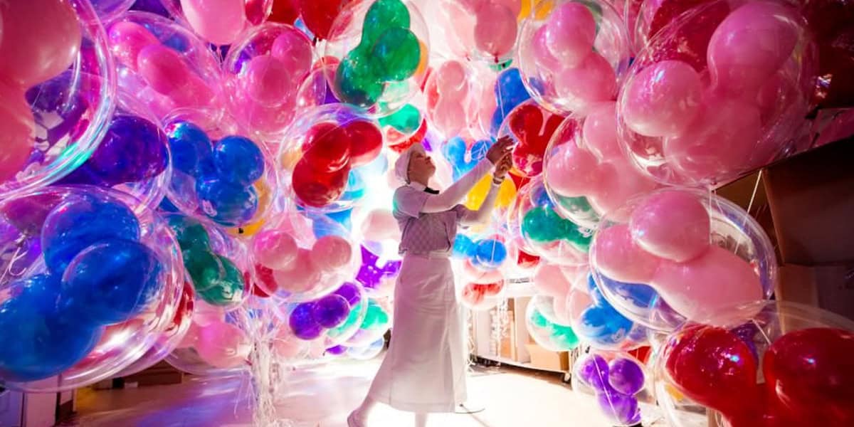 A person in white clothing stands surrounded by large, colorful balloons, creating a vibrant, immersive environment filled with Disney Magic and pink, red, blue, green, and purple hues.