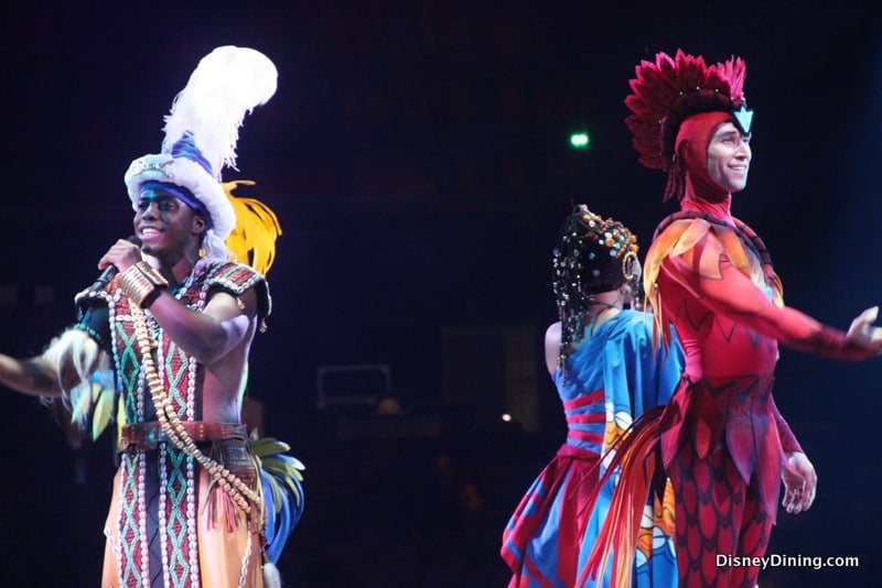 Disney performers in vibrant costumes sing and dance on stage, captivating the audience at a must-see theme park show.