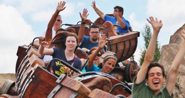 A group of people ride a wooden roller coaster, smiling and raising their hands in excitement as the train moves along the track under a partly cloudy sky.