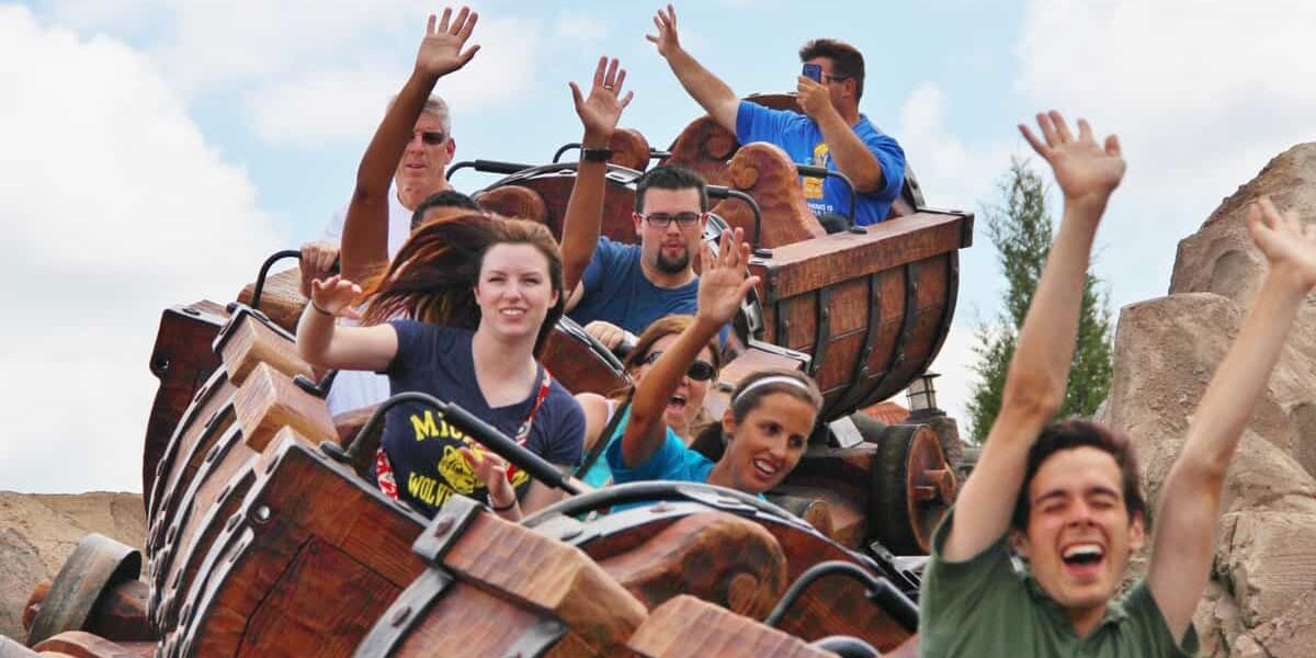 A group of people ride a wooden roller coaster, smiling and raising their hands in excitement as the train moves along the track under a partly cloudy sky.