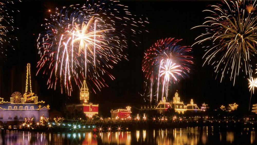 Vibrant fireworks dazzle over Disney World’s glowing attractions, their reflections shimmering in the calm lake beneath the night sky.