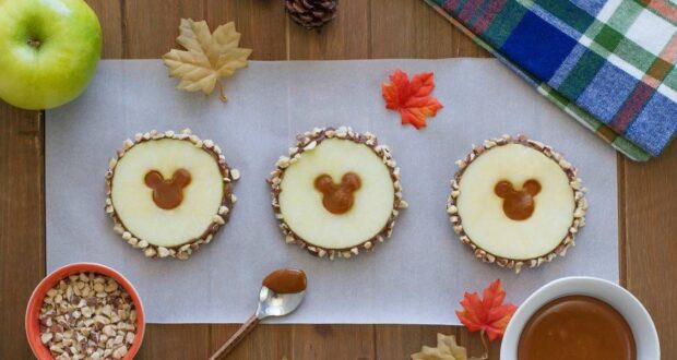 Three apple slices with caramel and nut coatings are arranged on parchment paper. Each slice has a Mickey Mouse-shaped caramel center. Surrounding the scene are leaves, an apple, nuts in a bowl, and a tartan-patterned napkin on a wooden surface.