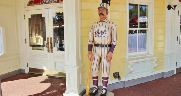A vintage baseball player statue with bat stands by a yellow Main Street Magic Kingdom building, evoking classic Disney nostalgia.