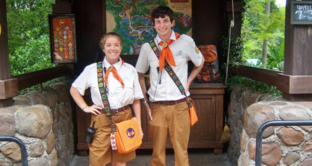 Two park employees in themed uniforms pose in front of a rustic kiosk. They wear khaki shorts, white shirts, orange scarves, and brown sashes. The background features a colorful map and stone details.