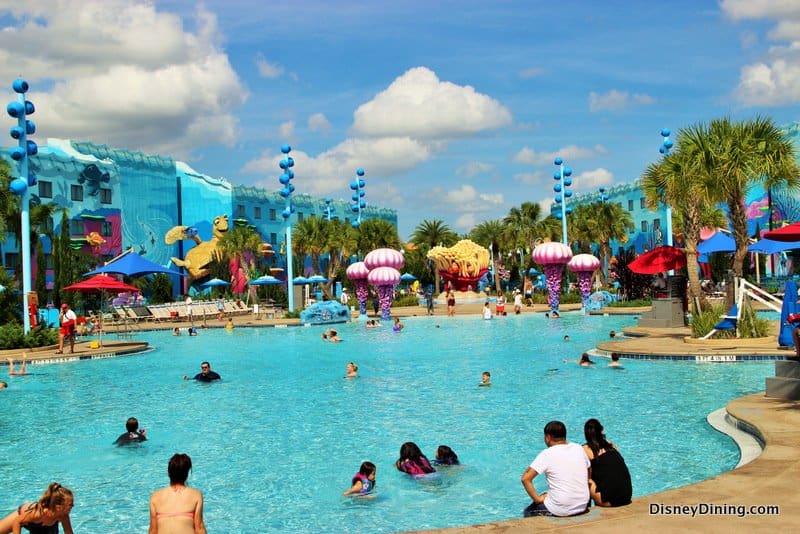 Guests enjoying a vibrant Disney resort pool, surrounded by colorful buildings and palm trees under the sunny Florida sky.