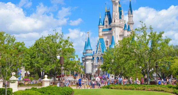 A large group of people walk and gather near Cinderella Castle at Disney World, surrounded by green trees and gardens under a bright, partly cloudy sky.