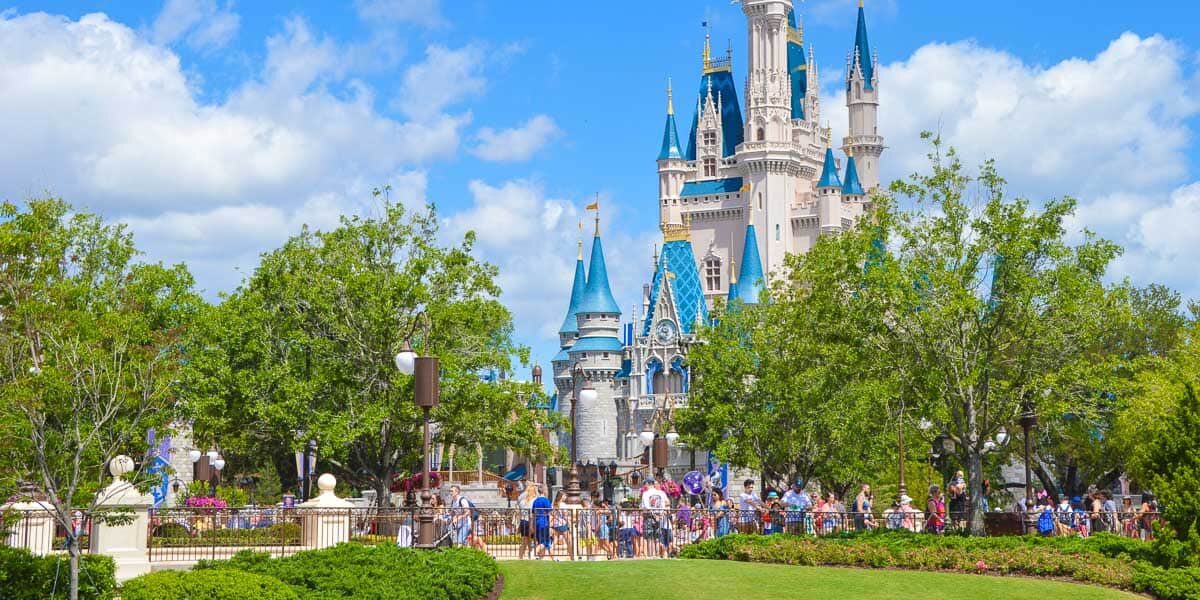 A large group of people walk and gather near Cinderella Castle at Disney World, surrounded by green trees and gardens under a bright, partly cloudy sky.