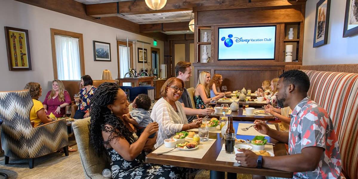 A group of people of diverse ages and backgrounds enjoy a meal together at a lively restaurant. They laugh and converse around wooden tables, with Disney Vacation Club signage and a Disney Vacation Club Guide visible in the background.