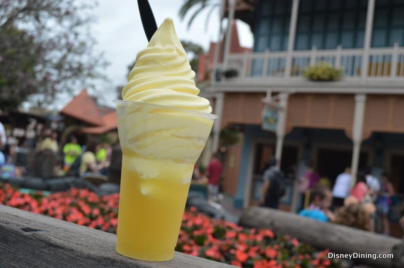 Dole Whip pineapple soft serve swirled in a clear cup with black spoon, served outdoors near an iconic Disney World building.