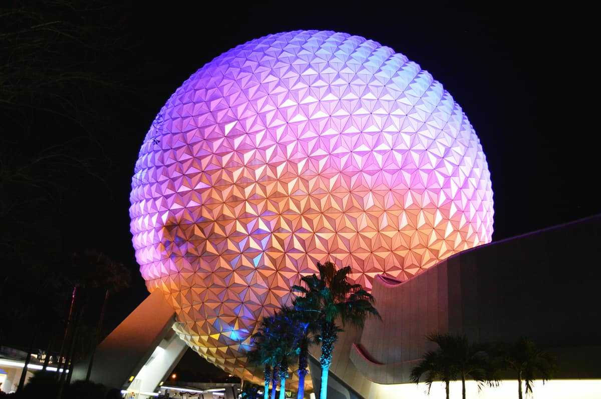 Spaceship Earth glows in vibrant purple, pink, and orange at night amid palm trees—a classic Epcot nighttime photo spot.