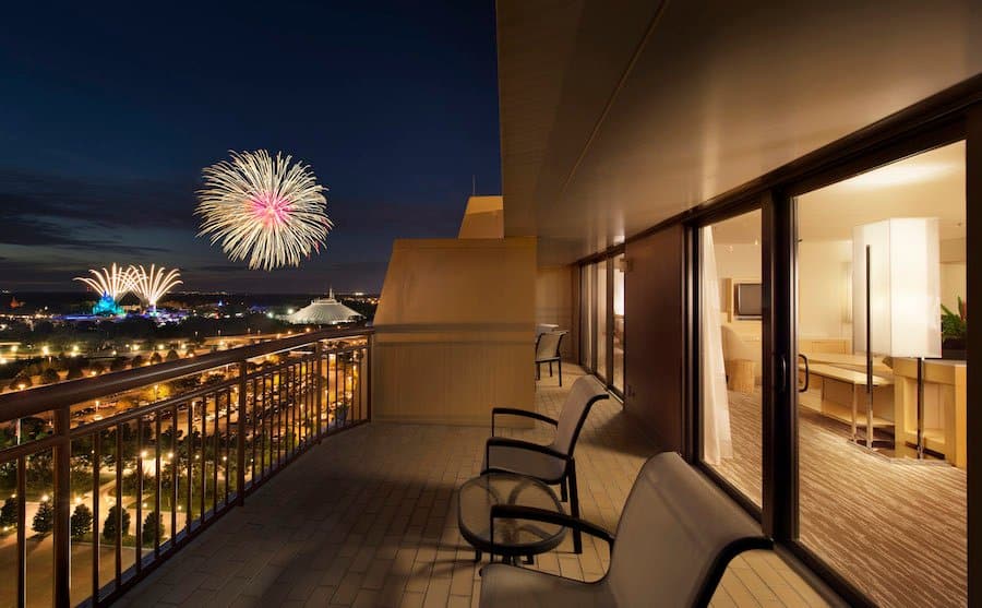 Resort balcony overlooking the city at night, with empty lounge chairs and fireworks lighting up the skyline like a theme park show.