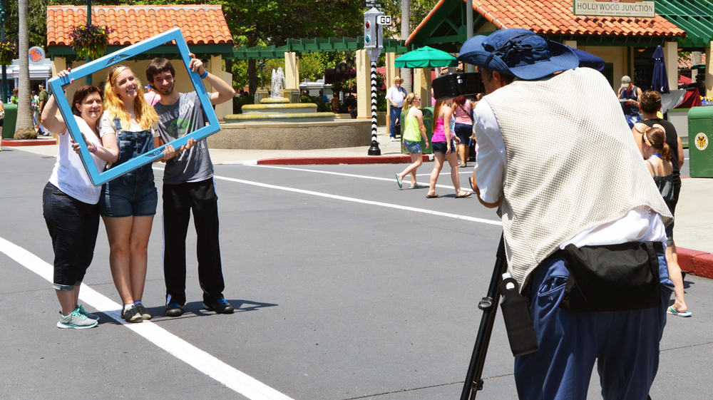 Three friends smile inside a blue photo frame as a family captures the excitement of Main Street U.S.A. on a bright day.