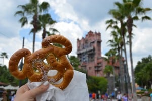 Mickey Mouse pretzel held up in front of Tower of Terror at Disney World, palm trees framing this classic theme park snack moment.