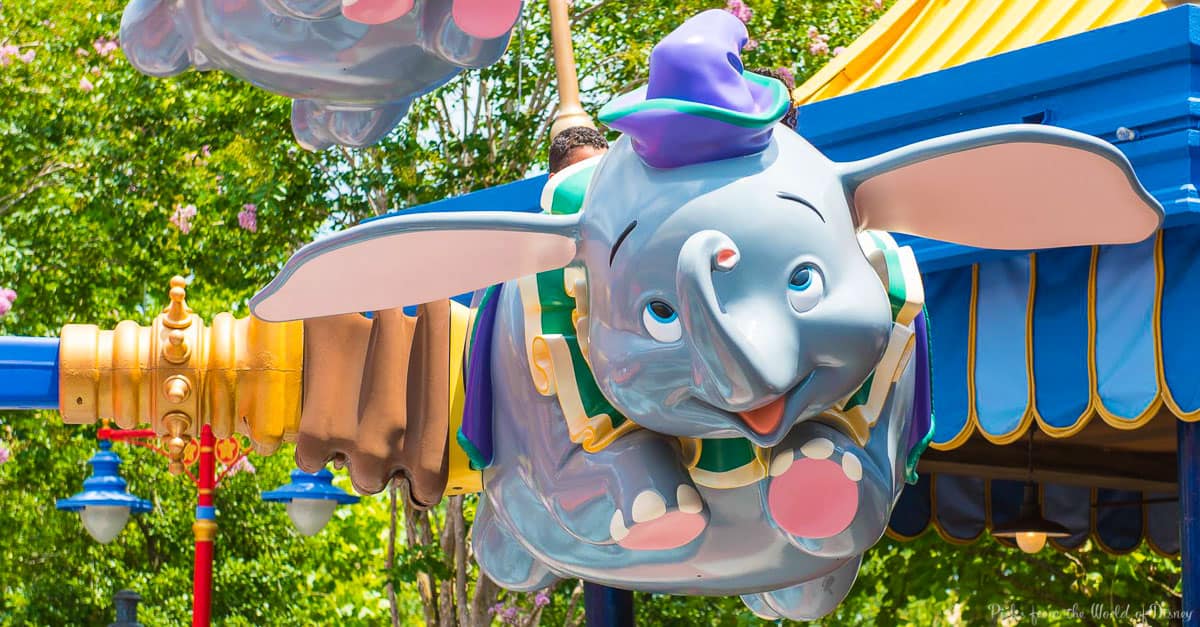 Colorful Dumbo ride at an amusement park, featuring a flying elephant ride vehicle with large ears and a hat, set against a sunny backdrop of trees and blue and yellow structures.