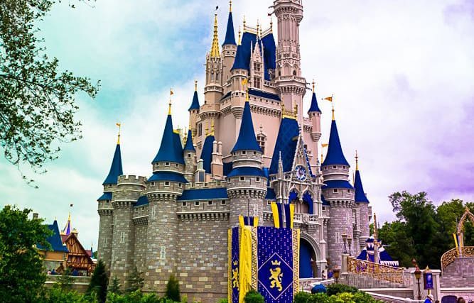The iconic Disney castle with blue spires and waving banners stands tall amid lush trees under a partly cloudy sky—pure magic.