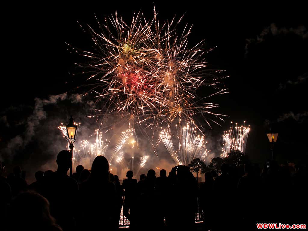 Guests at Magic Kingdom marvel as vibrant fireworks burst over Main Street, framed by glowing lamplights and leafy treetops.