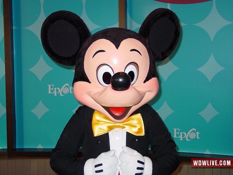 Mickey Mouse in a tuxedo and yellow bow tie greets guests against an Epcot backdrop, ready for your magical Disney World adventure.