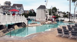 Disney’s resort pool with a scenic bridge, rows of empty loungers, lifeguard stands, and coastal-themed buildings in view.