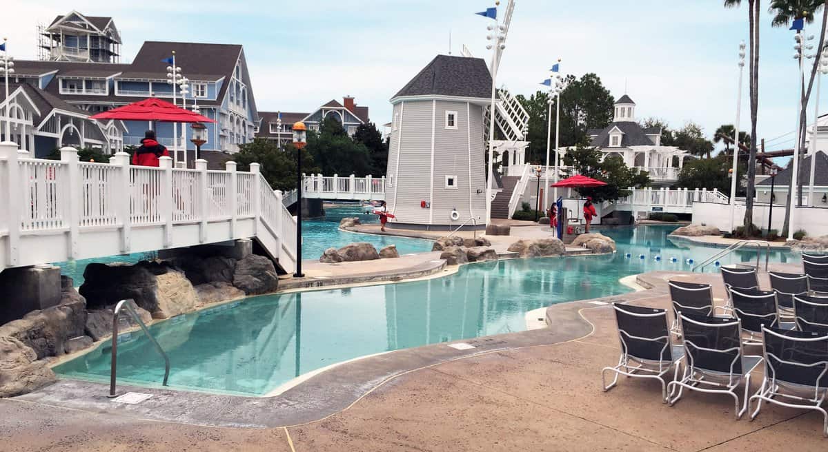 Disney’s resort pool with a scenic bridge, rows of empty loungers, lifeguard stands, and coastal-themed buildings in view.