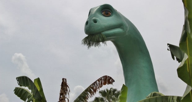 Gertie the Dinosaur statue munching foliage, surrounded by palms beneath cloudy skies at Disney’s Hollywood Studios.