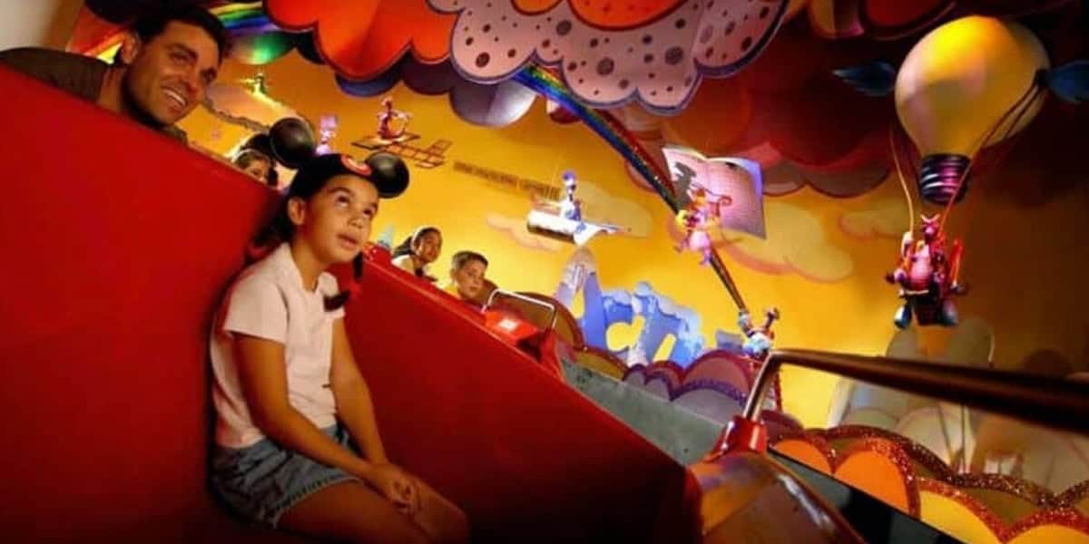 A young girl wearing Mickey Mouse ears and a man ride in a vibrant, whimsical amusement park attraction featuring colorful decorations, playful characters, hot air balloons, and catchy Disney songs, creating a joyful and animated atmosphere.