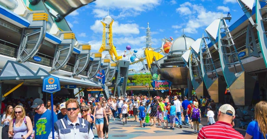 A large crowd of people walking through Tomorrowland at a theme park on a sunny day, surrounded by futuristic buildings and colorful signs under a blue sky with scattered clouds.