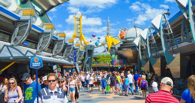 A large crowd of people walking through Tomorrowland at a theme park on a sunny day, surrounded by futuristic buildings and colorful signs under a blue sky with scattered clouds.