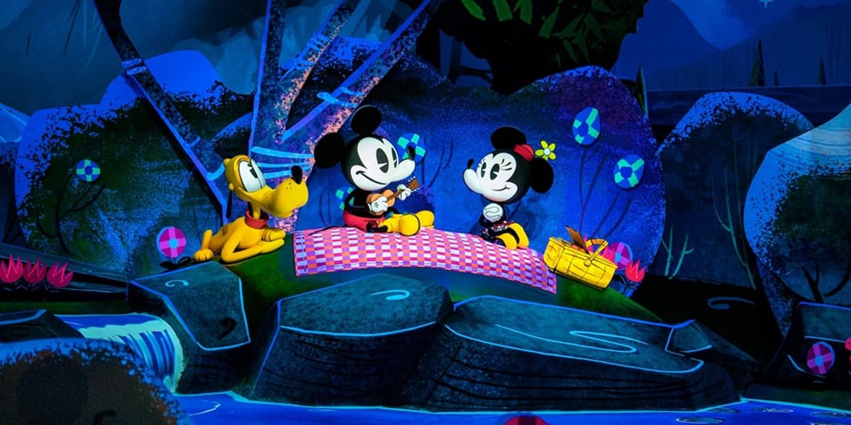 Mickey Mouse and Minnie Mouse enjoy a picnic on a red checkered blanket beside cheerful Pluto, surrounded by bright, cartoon-style scenery with flowers, a basket, and a waterfall as catchy Disney songs play in the background.