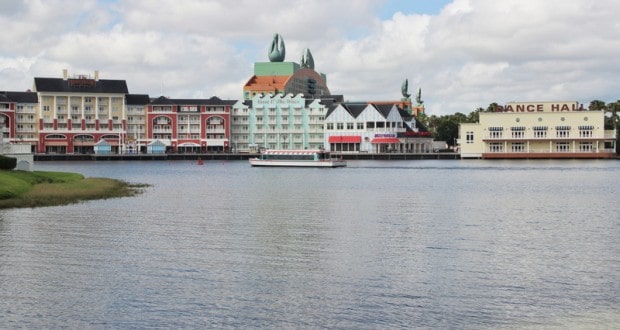 A Friendship Boat cruises Disney’s lake, passing vibrant resort facades and the lively Boardwalk Dance Hall under dramatic clouds.