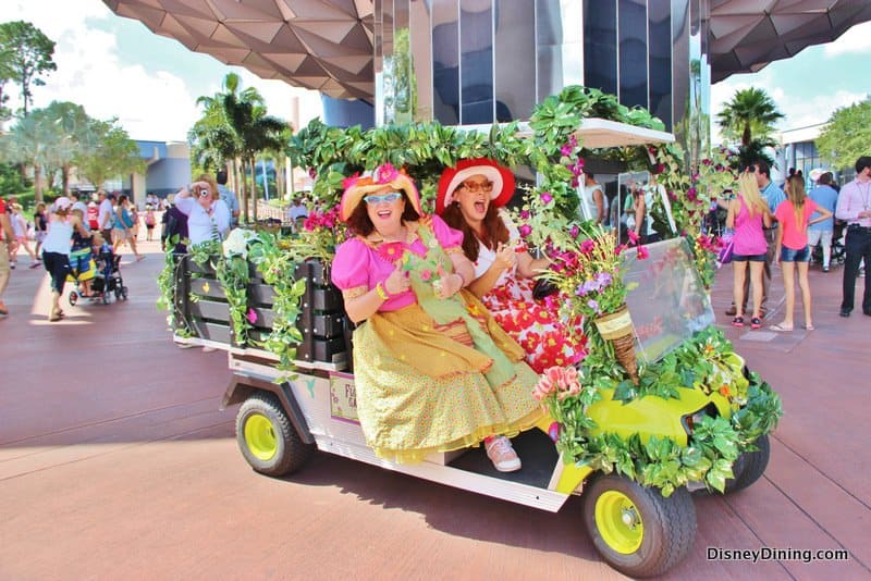 Cast Members in vibrant floral costumes and hats cruise Epcot in a flower-adorned golf cart, capturing Disney magic at work.