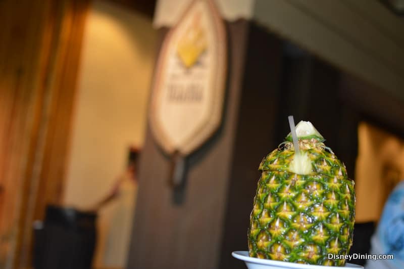 A pineapple drink with a straw on a plate, served at Tambu Lounge, with the restaurant sign blurred in the Disney World backdrop.