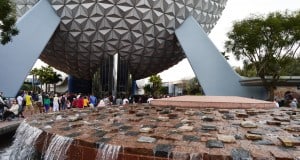 A futuristic, spherical building with a geometric pattern towers over a crowd of visitors near a red stone fountain with cascading water and trees in the foreground.