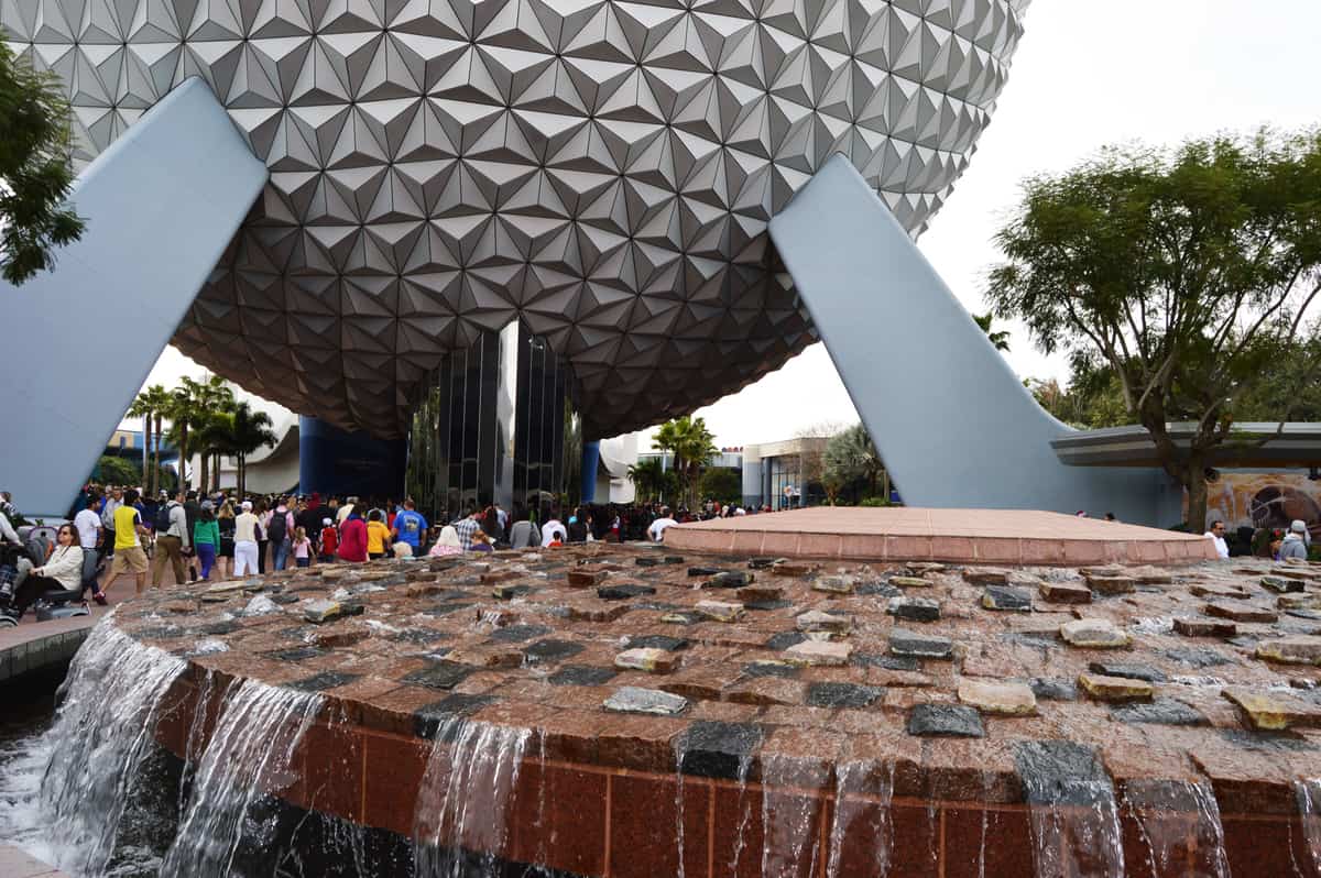A futuristic, spherical building with a geometric pattern towers over a crowd of visitors near a red stone fountain with cascading water and trees in the foreground.