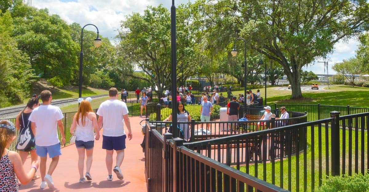 Guests strolling and mingling in a sunlit park at Disney World, surrounded by trees, grass, and a charming fenced walkway.