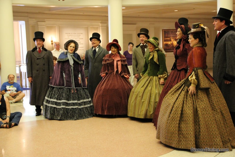 Victorian carolers perform inside The American Adventure at Epcot, entertaining casually dressed guests gathered around.